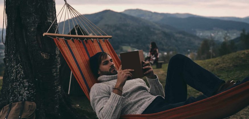 Leave all your worries behind. Handsome young man lying in hammock and reading a book while camping with his girlfriend