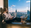Blonde hair caucasian female student closed her eyes while enjoying warm evening sunset light though hotel window with panoramic New York city views. Hipster girl traveler working on laptop computer