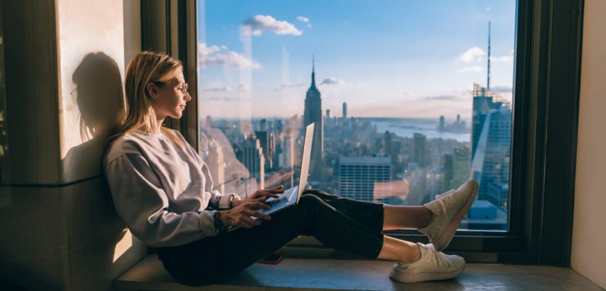 Blonde hair caucasian female student closed her eyes while enjoying warm evening sunset light though hotel window with panoramic New York city views. Hipster girl traveler working on laptop computer