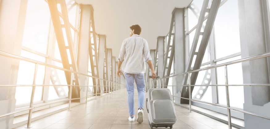 Rear View Of Young Man Walking With Luggage In Airport Terminal